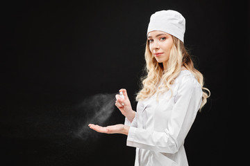 Young woman in medical uniform using hand sanitizer spray, isolated at black background. Hand hygiene and antibacterial protection.