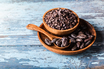 Organic raw cocoa beans, nibs in wooden bowls on blue old rustic background.