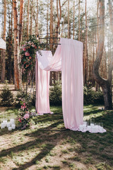 the arch for the wedding ceremony is decorated with floral arrangements and pink fabric and stands in a green meadow against the background of the forest