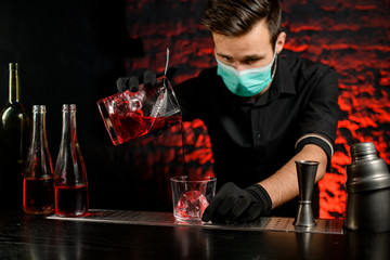 Professional bartender with mask pours cold cocktail into glass.