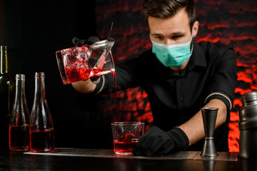 Barman with mask masterfully pours cold cocktail into glass.