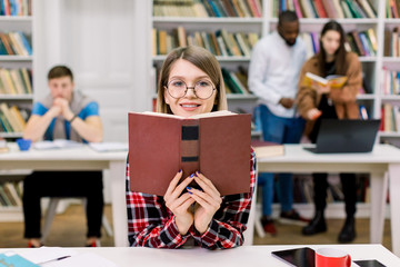 Portrait of charming smiling student girl in casual wear and eyeglasses studying at library, sitting at the table and holding open book near her face. Mixed race friends atudying on the background