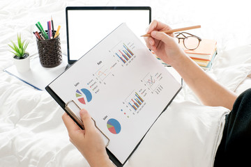 Young woman hand holding document graph with pencil and sitting in the bed room, Work form home and holiday concept with document graft and laptop computer, supplies