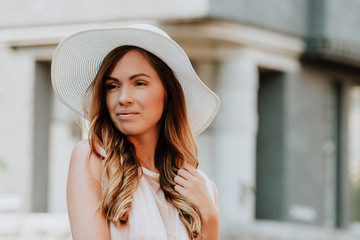 Outdoor close up fashion portrait of young beautiful happy smiling lady wearing white dress, stylish straw wide brim hat, posing on street. City