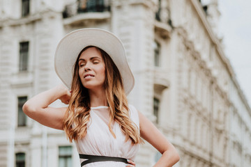 Outdoor fashion portrait of young beautiful happy smiling lady wearing white dress, stylish straw wide brim hat, posing on street. City