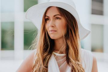 Outdoor fashion portrait of young beautiful happy smiling lady wearing white dress, stylish straw wide brim hat, with elegant handbag,posing on street. City