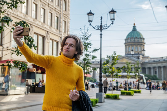 The handsome men does selfie, Look from outside, he dressed in a yellow sweater, a black raincoat or jacket is his hands, Bolshaya Konyushennaya street and Kazan Cathedral on background, sunny day