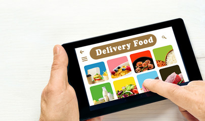 Male hands holding tablet computer with app delivery food on screen above white desk.