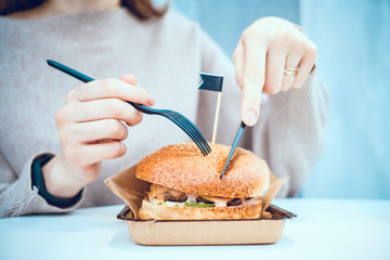 Burger in craft paper on the table and a girl eating his fork and knife.