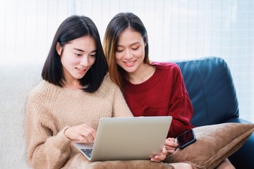 Young beautiful Asian women lesbian couple lover using smartphone and laptop video call online in living room at home with smiling face.Concept of LGBT sexuality with happy lifestyle together.