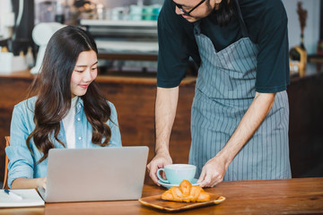 Young beautiful Asian woman working with laptop and barista holding hot coffee cup in coffee shop background and .Concept of female freelancer.Smiling and happy face with vintage tone