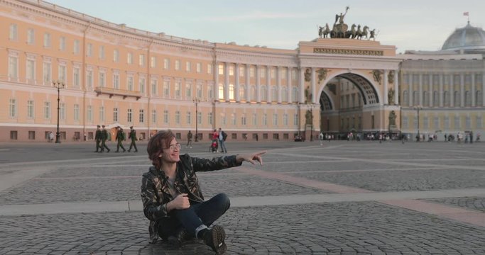 The beautiful young man siting on the Palace square and drinks coffee at sunset, he dressed in a military jacket and jeans, long curly hairs, Arch of the General Staff Building is on background