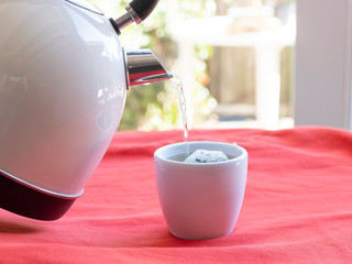 Electric kettle with boiling water for the herbal tea. Tea bag in a white tea cup on a red table, Selective focused on tea cup