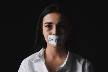 Scared female victim with taped mouth on dark background. Concept of violence