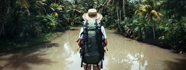 Traveling woman with backpack and straw hat looking at tropical river at sunny day. Wide screen...
