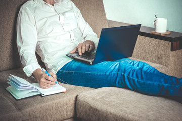 Naklejka premium young man in white shirt and blue jeans working on laptop, writes in notebook in home office sitting on cozy beige sofa Stay at home, online work, distance learning concept