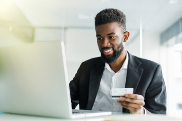 Portrait of confident young african man holding credit card with laptop paying via internet