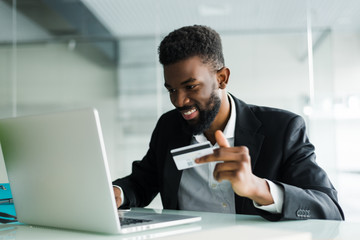 Portrait of confident young african man holding credit card with laptop paying via internet