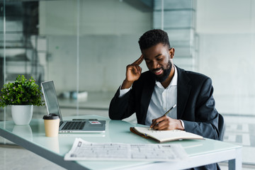 Young african businessman working in office at laptop and make notice in notebook
