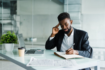 Young african businessman working in office at laptop and make notice in notebook