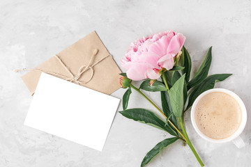 Morning coffee cup, pink peony flower and blank greeting card with envelope on light background....