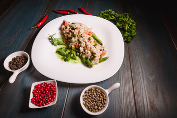 Appetizing risotto on a white grater among seasonings, stands on a wooden table. Studio photography of food in the cooking industry, dark background