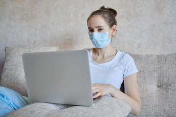 A girl in a medical mask is in quarantine at home working on a laptop.