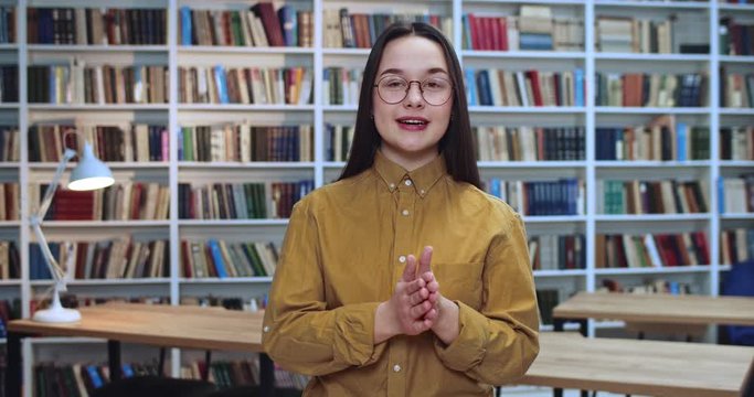 Portrait of nice beautifull geek student in glasses emotionally explaining and making a lot of gests by her hands in library.