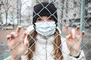 The girl in the medical mask is in the street in isolation and is holding fence.