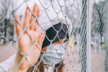 A girl in a medical mask is isolated on the street and leaned on a fence grate.