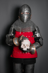 A medieval knight in full armor holds in his hands a human skull standing on a gray background.