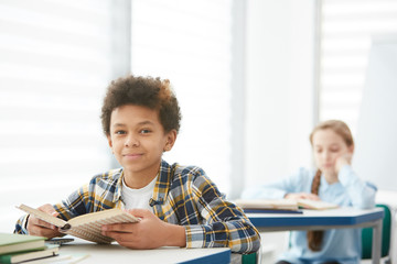 Portrait of smiling African-American boy sitting at desk in school and looking at camera while...