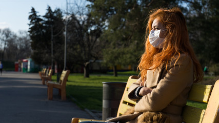 Sad beautiful red haired girl sitting in empty park wearing surgery mask due to pandemia.Woman in...