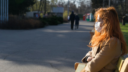 Sad beautiful red haired girl sitting in empty park wearing surgery mask due to pandemia.Woman in mask outdoor  caused by epidemic in city.Virus protection.