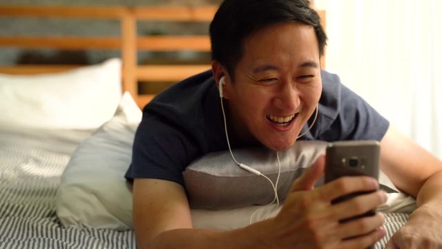 Portrait of happy 30s Asian man in casual clothing making facetime video calling with smartphone at home, waving at people on phone screen. Using conferencing meeting online app, social distancing