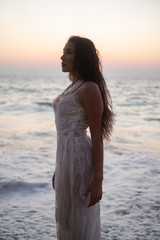 Romantic portrait of beautiful brunette woman in white lace dress standing at the beach over sea and sunset sky background