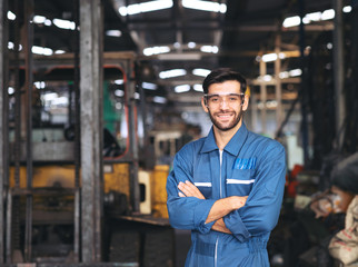 Smiling worker in safety glassess standing with arm crossed near industrial equipment manufacturing...