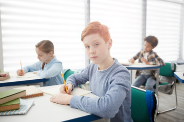 Obraz premium Portrait of modern red-haired schoolboy sitting at desk in classroom and looking at camera while taking notes, copy space