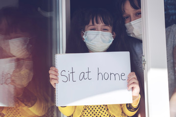 Two girls in medical masks look out of a window with an album labeled "Sit at home". Little girls in quarantine for coronavirus