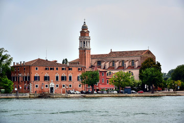 Venice, Italy - A brown building with a tiled roof of the church and monastery of San Nicolo del Lido, green trees on the shore near the water, gray sky in the summer in the daytime.