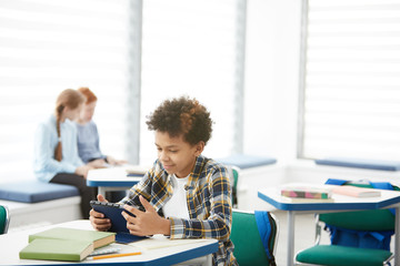 Portrait of smiling African-American boy using digital tablet while sitting at desk in school classroom, copy space