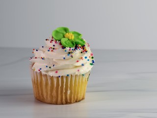 Yellow cupcake with white swirled frosting coated in sprinkles and topped with a green fondant flower as a decoration, sitting on a sandstone counter with a white background.  Festive, celebration.