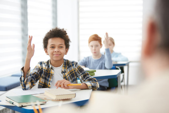 Portrait of smiling African-American boy raising hand to answer while sitting at desk in school classroom, copy space