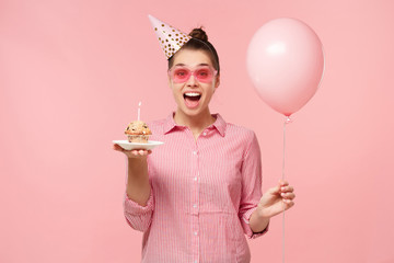 Excited birhday girl celebrating, holding air balloon and cake,  isolated on pastel pink studio...
