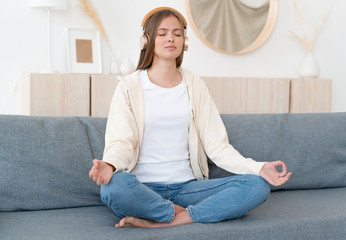 Fototapeta premium Calm young woman sitting on couch, practicing meditation yoga with mudra hands, listening to relaxing peaceful zen music in wireless headphones