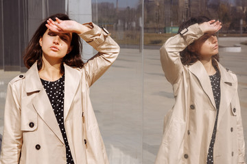 A young beautiful girl stands next to a mirrored building and holds her head. Headache. Reflection in the mirror. Girl in a raincoat and dress in peas.