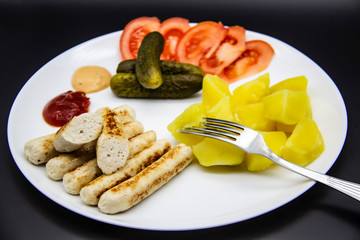 Delicious homemade chicken sausages with tomatoes, cucumbers, potato and souces. Fragrant sausages made from non-GMO chicken. Toasted Mini Sausages from chicken on the plate. Ukrainian tasty food.