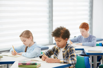 Multi-ethnic group of kids sitting at desks in school classroom and writing or taking test, copy space - obrazy, fototapety, plakaty