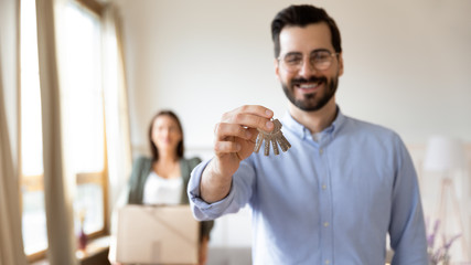Fototapeta premium Head shot focus on happy young husband in glasses demonstrating keys to camera, while smiling wife carrying cardboard boxes. Excited married family couple celebrating moving day in new apartment.