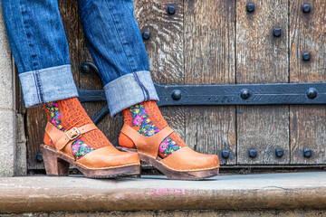 A woman's legs wearing wooden clog shoes with brown leather in front of an old wooden vintage door